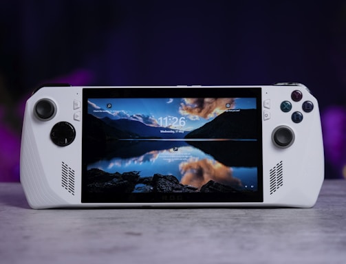 A handheld gaming console with a central screen displaying a scenic wallpaper of mountains and a lake, flanked by controller buttons and joysticks on both sides. The lighting around is dim, with purplish hues in the background, creating a modern and elegant atmosphere.